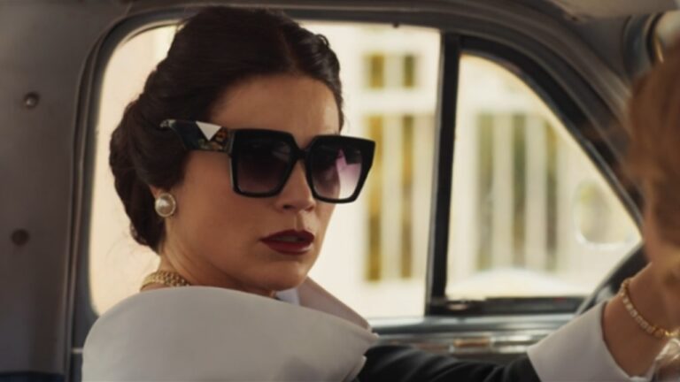 Mulher elegante dentro de um carro antigo, usando óculos de sol grandes, brincos de pérola e batom vermelho. Ela olha séria para o lado enquanto segura o volante, transmitindo postura firme e sofisticada.