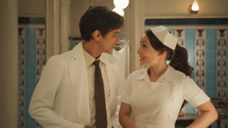 Cena mostra um médico e uma enfermeira conversando animadamente em um ambiente hospitalar de época, ambos usando uniformes brancos; eles sorriem um para o outro diante de uma parede com azulejos decorativos azuis e vermelhos.