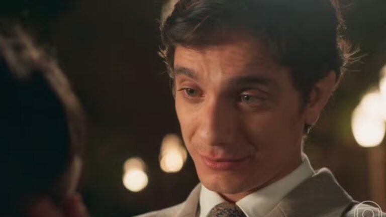 Homem jovem em close com pele clara cabelo castanho curto penteado de lado vestindo terno claro camisa branca e gravata clara com expressão suave e levemente emotiva olhando para outra pessoa fora de quadro à esquerda, iluminação quente e baixa destaca o rosto e cria reflexos nos olhos transmitindo clima íntimo e romântico, ao fundo aparecem luzes desfocadas em tons dourados sugerindo ambiente noturno externo ou jardim iluminado, enquadramento focado no rosto e parte do tronco reforça a sensação de conversa próxima e momento emocional, no canto inferior direito há um pequeno logotipo de emissora indicando cena de produção televisiva