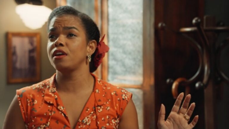 Uma mulher aparece em ambiente interno com iluminação quente, falando com expressão séria e levemente emocionada. Ela tem pele negra, cabelos presos para trás com um acessório vermelho e usa um vestido sem mangas de cor laranja com estampa floral. A mão direita está levantada, como em gesto de explicação ou apelo. Ao fundo, há uma janela com vidro fosco, um cabideiro de madeira e um quadro na parede, sugerindo uma cena de época ou ambiente doméstico antigo, possivelmente de filme ou novela.