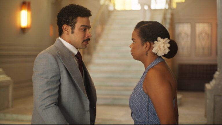 A imagem mostra um homem e uma mulher em uma conversa séria dentro de um prédio elegante, com uma escadaria ao fundo. O homem veste um terno cinza e gravata escura, enquanto a mulher usa um vestido azul com bolinhas e tem o cabelo preso em um coque adornado com uma flor branca. A cena parece ser de época, transmitindo um tom dramático.