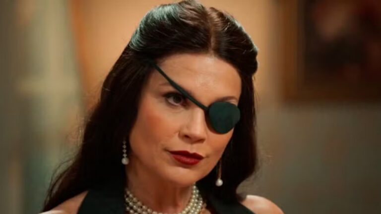 Mulher com tapa-olho preto, colar de pérolas e vestido escuro em cena de novela com expressão séria