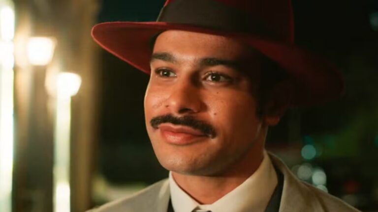 Homem com chapéu vermelho e bigode sorri levemente em cena noturna de época com luzes desfocadas ao fundo