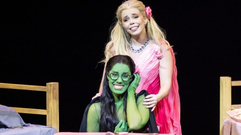 Atrizes caracterizadas como Elphaba e Glinda em cena do musical Wicked com figurinos verde e rosa em palco escuro