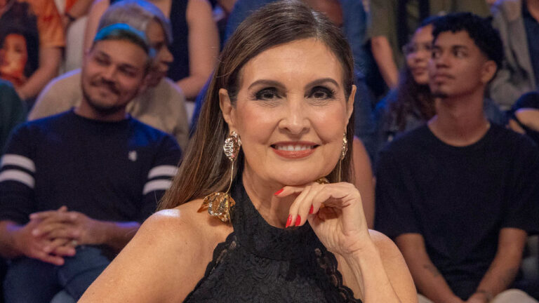 Fátima Bernardes, em um plano fechado, sorrindo e apoiando o queixo em uma das mãos com as unhas pintadas de vermelho, usando um vestido preto de renda no ombro e brincos longos e dourados com um design floral. Ela tem cabelos castanhos escuros e está em um estúdio com uma plateia ao fundo.