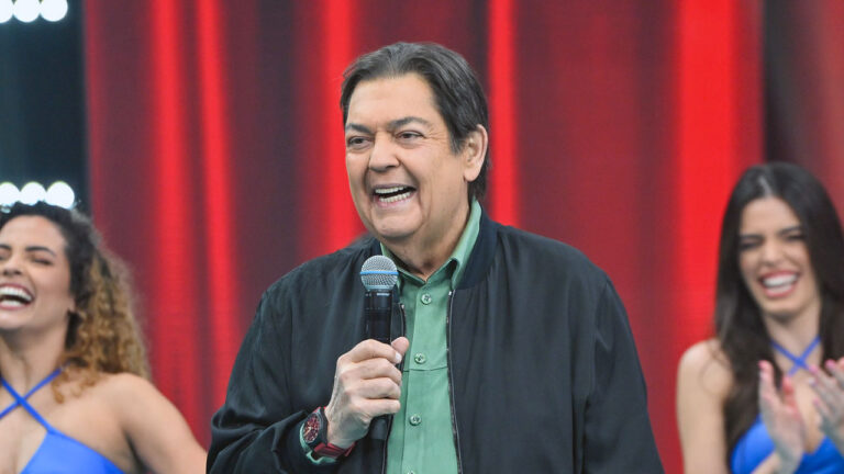 Fausto Silva sorri enquanto segura um microfone no palco do Faustão na Band com dançarinas e fundo vermelho ao fundo no estúdio