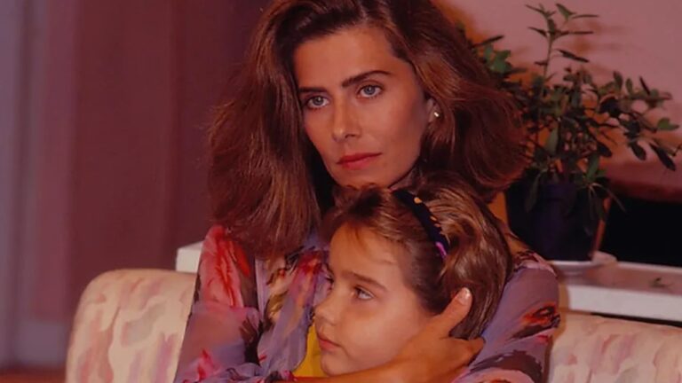 Mulher de expressão séria abraça uma menina que está sentada em seu colo em ambiente interno com planta em vaso ao fundo