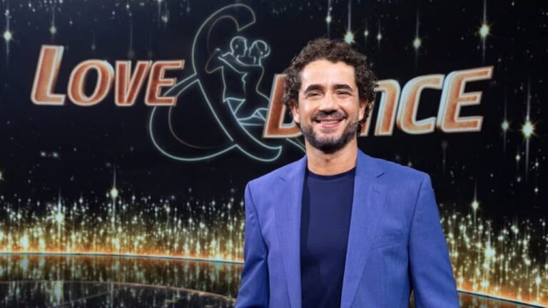 Homem adulto em pé sorrindo para a câmera com cabelo cacheado escuro e barba curta usando blazer azul e camiseta azul escura diante de cenário de estúdio com fundo preto iluminado por pontos dourados e o logotipo Love and Dance ao fundo com desenho estilizado de casal dançando e letras grandes iluminadas sugerindo programa de televisão ou evento de entretenimento
