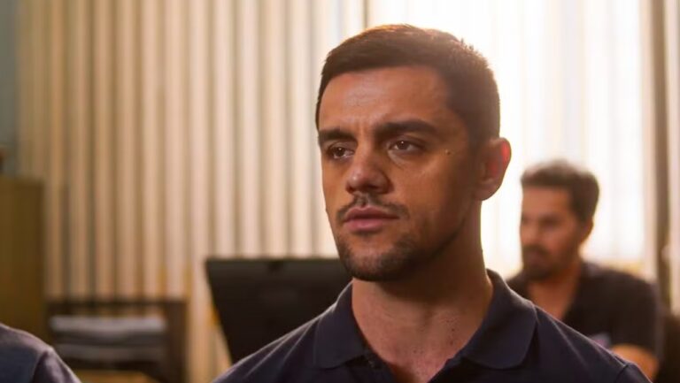 Homem em primeiro plano, com expressão séria, em um ambiente interno iluminado por luz quente. Ao fundo, outra pessoa aparece desfocada, sugerindo uma cena de conversa ou tensão em um espaço fechado.