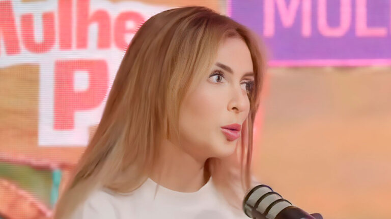 Fernanda Campos de blusa branca fala ao microfone olhando para o lado durante entrevista em podcast com painel rosa e violeta