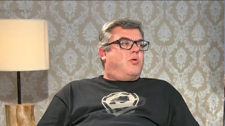 Homem de meia-idade usando óculos e camiseta escura, sentado em ambiente interno com papel de parede estampado e luminária ao fundo, fala enquanto olha para o lado, em uma cena de entrevista ou conversa.