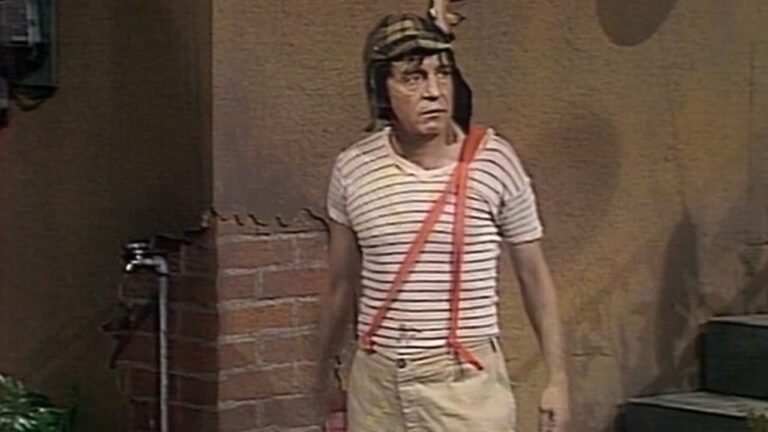 A imagem mostra o personagem Chaves, da clássica série de comédia mexicana. Ele está no pátio da vila, vestindo sua tradicional camiseta listrada, calças bege com suspensórios vermelhos e um gorro com abas xadrez. A cena apresenta o cenário simples e característico do programa, com parede de tijolos e torneira ao fundo