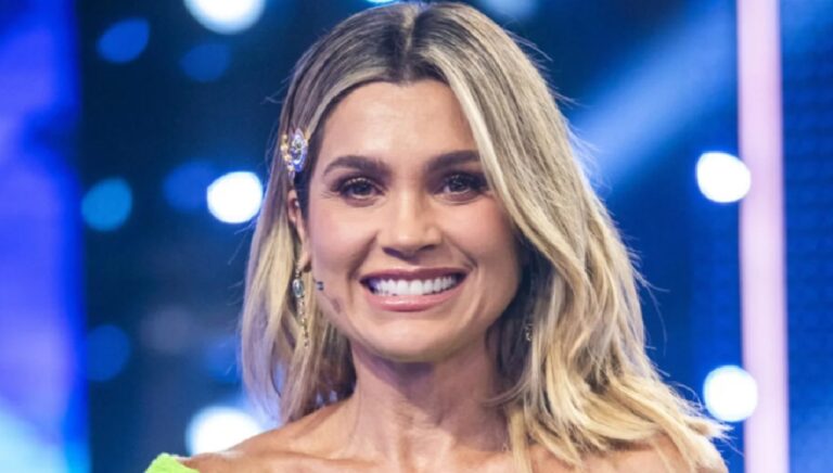 Mulher adulta sorri para a câmera em um estúdio de televisão iluminado por luzes azuis. Ela tem cabelos loiros soltos, usa um acessório brilhante no cabelo e maquiagem discreta. O fundo desfocado com pontos de luz sugere um cenário de programa ou evento televisivo.