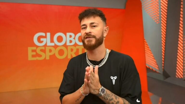 Apresentador em estúdio do Globo Esporte, em frente a um painel laranja com o nome do programa ao fundo. Ele aparece de mãos unidas, usando camiseta preta, correntes prateadas e exibindo tatuagens nos braços, em um cenário iluminado com elementos geométricos modernos.
