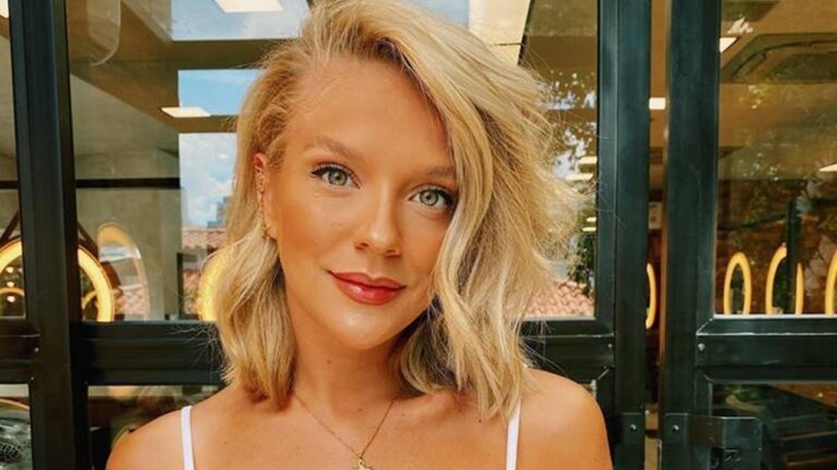 Mulher adulta de cabelos loiros curtos e ondulados posa para a câmera em ambiente interno bem iluminado. Ela tem olhos claros, maquiagem discreta e expressão serena. Usa blusa clara de alças finas e colar delicado. Ao fundo, há portas de vidro com reflexos de luz e elementos arquitetônicos modernos, sugerindo um espaço comercial ou salão.