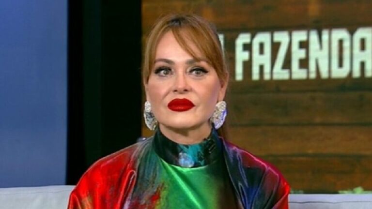 Mulher sentada em estúdio de televisão, usando brincos grandes e um vestido brilhante com cores vibrantes. Ela está maquiada com batom vermelho intenso e olha séria para a frente. Ao fundo, aparece um painel com letras parcialmente visíveis.