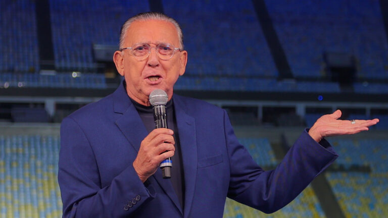 Galvão Bueno de terno azul e óculos fala ao microfone e gesticula com a mão em estádio com arquibancadas azuis ao fundo