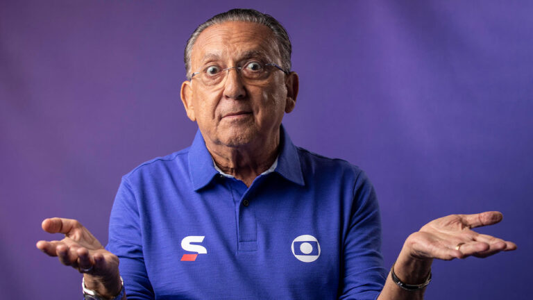 Galvão Bueno de camisa polo azul da Globo abre os braços com expressão de dúvida em foto promocional com fundo roxo da emissora