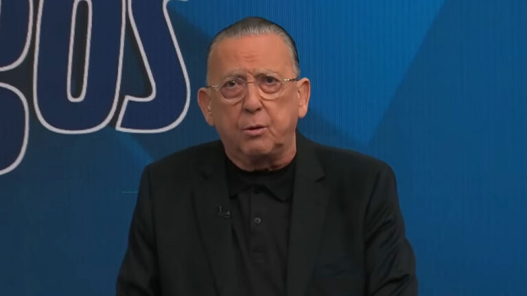 Galvão Bueno usa paletó preto e óculos redondos enquanto fala no cenário azul do programa Galvão e Amigos na Band