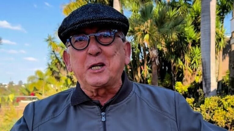 Homem idoso usa boina escura, óculos e jaqueta cinza enquanto fala ou sorri em ambiente externo com árvores e céu azul ao fundo