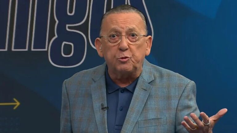 Homem de óculos e terno azul fala gesticulando em estúdio de programa esportivo com fundo azul escuro