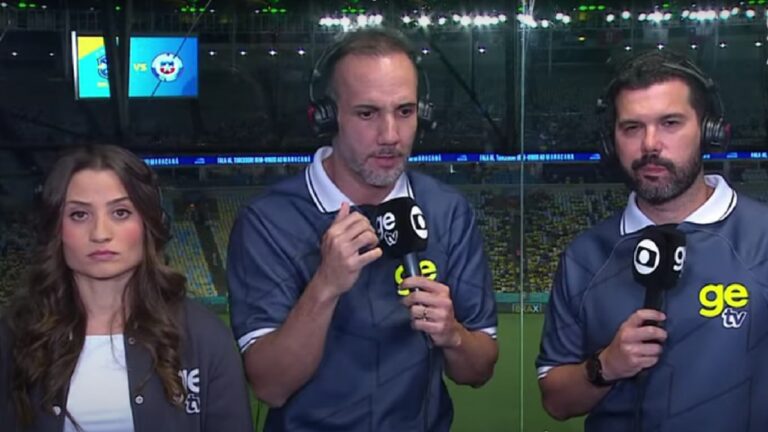Três comentaristas esportivos estão em cabine de transmissão de estádio durante uma partida. Eles usam fones de ouvido e camisas azul-escuro com o logo “ge tv”. A mulher à esquerda mantém expressão séria; o homem ao centro fala segurando um microfone; o homem à direita também segura um microfone. Ao fundo, vê-se o campo iluminado e um telão exibindo escudos dos times em jogo.