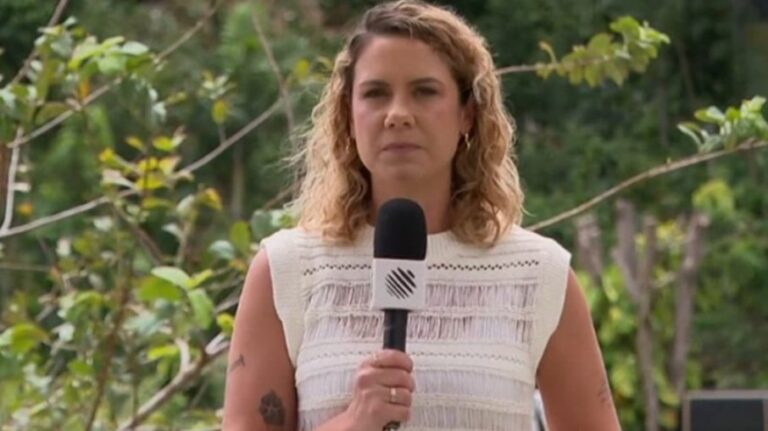 Repórter segura um microfone com o logotipo de um canal de TV enquanto faz uma entrada ao vivo ao ar livre; ela usa blusa branca sem mangas e aparece diante de vegetação verde, com expressão séria e postura firme.