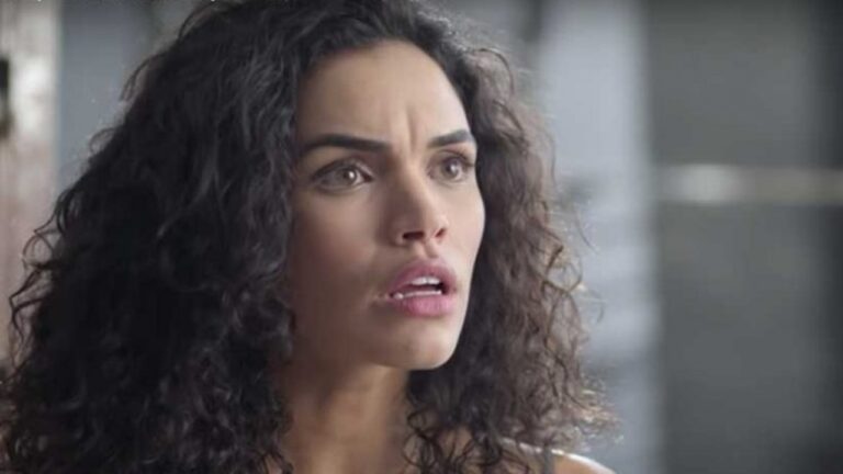 Mulher de cabelos cacheados escuros aparece em close, com expressão de surpresa e preocupação; seus olhos estão arregalados e a boca entreaberta, enquanto o fundo desfocado sugere um ambiente interno iluminado suavemente