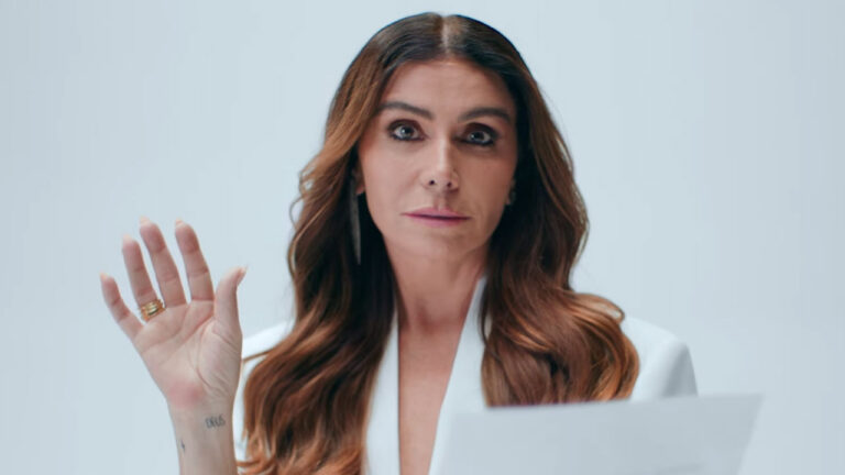 Giovanna Antonelli com cabelo solto e blazer branco ergue a mão com expressão séria ao segurar papel em estúdio de fundo claro na campanha da HBO Max