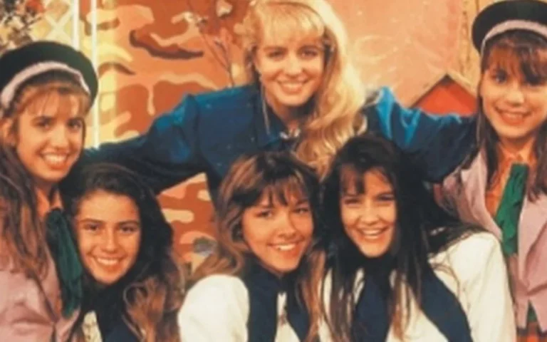 A foto parece antiga (provavelmente anos 1980 ou início dos 1990), com seis jovens e uma mulher adulta ao centro, todos sorrindo e muito próximos, passando uma sensação de grupo unido. Eles usam roupas parecidas, com lenços no pescoço e alguns chapéus, o que sugere um elenco infantil/juvenil, possivelmente de um programa de TV, grupo musical ou projeto artístico da época. A mulher ao centro parece ter uma posição de liderança ou destaque, com os braços abertos sobre os jovens.