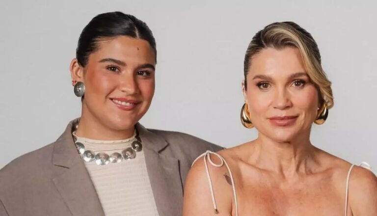 Duas mulheres posam lado a lado diante de fundo neutro. A mulher à esquerda veste blazer bege, blusa clara e colar metálico, sorrindo suavemente. A mulher à direita usa vestido de alças finas e brincos dourados grandes, olhando para a câmera com expressão serena