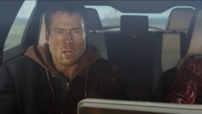 Homem com o rosto sujo e expressão de choque está sentado no banco traseiro de um carro, olhando fixamente para algo à sua frente. Ele veste um moletom com capuz sob uma jaqueta escura. O interior do veículo é claro e moderno, com os bancos dianteiros parcialmente visíveis