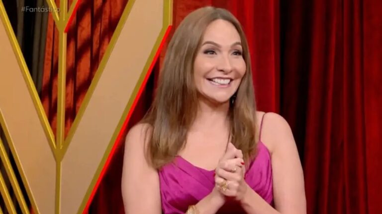 Mulher sorridente com cabelo liso castanho claro veste vestido rosa de alças finas e acessórios dourados, posando diante de um fundo com cortina vermelha e estrutura dourada iluminada com detalhes em vermelho, com a palavra “#Fantástico” no canto superior esquerdo da imagem