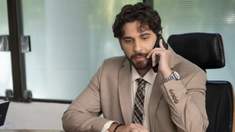 Homem jovem sentado à mesa de escritório, usando terno bege e gravata estampada, enquanto segura um telefone celular na orelha. Ele parece concentrado na conversa, com o corpo levemente inclinado para frente. Ao fundo, janelas amplas com vidro fosco deixam entrar luz suave.