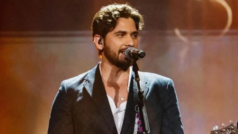 Homem canta em um palco, posicionado de frente para um microfone em pedestal, sob iluminação quente em tons alaranjados. Ele tem barba curta, cabelo escuro ondulado e usa um paletó escuro sobre camisa clara parcialmente aberta no colarinho. Um violão aparece parcialmente à direita do enquadramento, preso por uma correia sobre o ombro. A cena sugere uma apresentação musical ao vivo, com clima intimista e foco no intérprete.