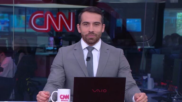 A imagem mostra Gustavo Uribe, apresentador de telejornal da CNN Brasil, sentado à bancada do estúdio, usando terno cinza, camisa branca e gravata. À sua frente há um notebook preto e uma caneca branca com o logotipo da emissora. O ambiente ao fundo é uma redação jornalística, com computadores e profissionais trabalhando