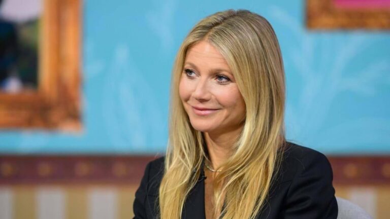 Mulher loira sentada em estúdio de entrevista sorri de forma discreta; ela usa blazer escuro e mantém os cabelos lisos soltos, com um fundo azul desfocado decorado por molduras coloridas.