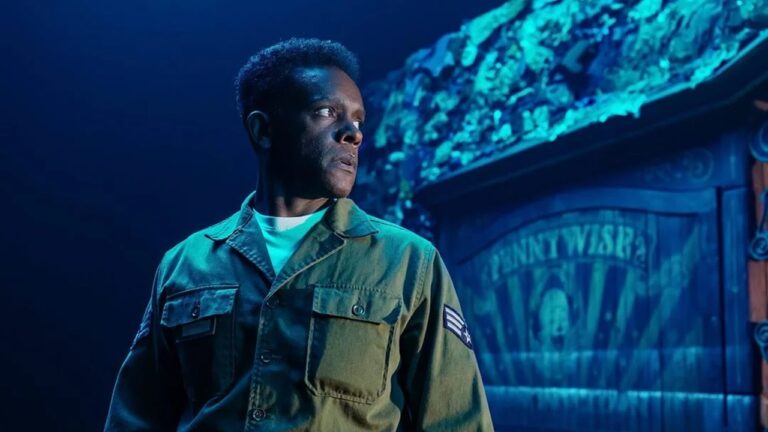 Homem negro usando uniforme militar verde-oliva está em pé sob iluminação azul dramática, olhando para a esquerda com expressão séria e atenta. Ao fundo, há uma construção de madeira com aparência rústica e iluminação azul-esverdeada, criando um clima noturno e cinematográfico. O cenário sugere uma situação de tensão ou vigilância, possivelmente em um ambiente fictício ou de filme.