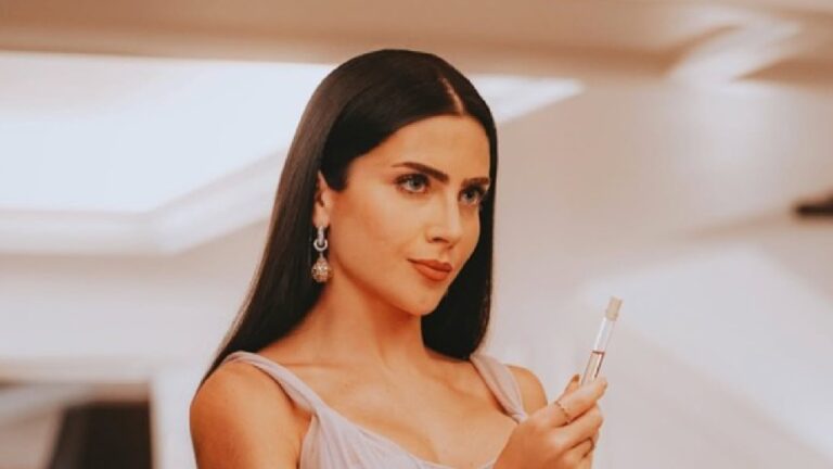 Mulher jovem de cabelos longos e lisos, vestindo um vestido leve em tom claro, segura um pequeno frasco transparente semelhante a um perfume ou cosmético. Ela olha para a frente com expressão confiante, usando brincos pendentes e posando em um ambiente interno bem iluminado