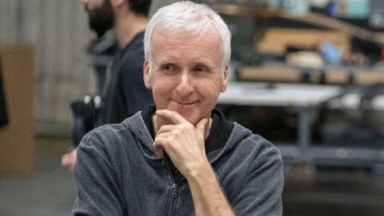 Homem adulto de cabelos curtos e grisalhos aparece em primeiro plano, sorrindo de forma discreta com a mão apoiada no queixo, em gesto pensativo. Ele veste um moletom escuro com zíper. O fundo está desfocado e mostra um ambiente interno amplo, com mesas e equipamentos, sugerindo um estúdio, oficina ou espaço de trabalho.