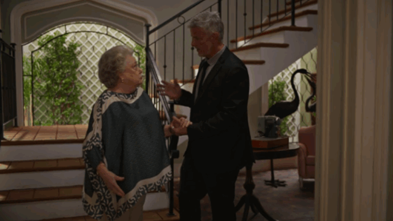 Uma mulher idosa e um homem de cabelos grisalhos conversam em um hall de entrada iluminado, próximo a uma escada com grades pretas. A mulher usa uma capa estampada em tons de verde e bege, enquanto o homem veste terno escuro e segura a mão dela. Ao fundo, há plantas, uma parede com treliça branca e parte de uma sala com móveis