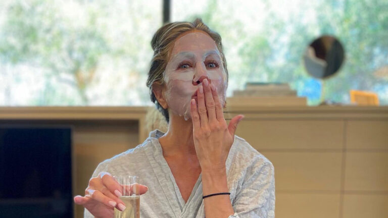 Jennifer Aniston com máscara facial branca manda beijo e segura taça de bebida em momento de cuidados com a pele e relaxamento