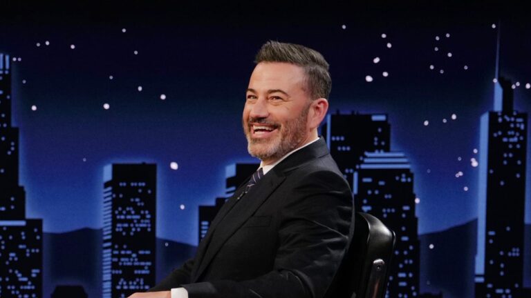 Apresentador sentado em estúdio noturno, sorrindo enquanto olha para o lado, com cenário ao fundo mostrando silhuetas de prédios iluminados contra um céu estrelado