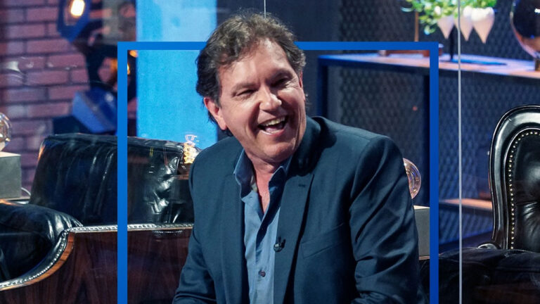 Empresário João Appolinário sorri abertamente, vestindo blazer azul escuro e camisa social, durante participação em programa de TV