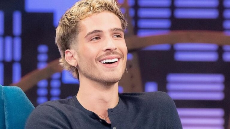 Um jovem sentado em um estúdio de TV sorri amplamente, com expressão descontraída. Ele usa uma camisa escura e tem cabelo curto com mechas claras. O fundo desfocado mostra luzes coloridas em tons de roxo e azul, reforçando o clima de programa televisivo.