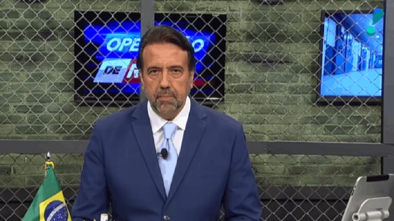 Apresentador de televisão em estúdio jornalístico, vestindo terno azul e gravata clara, olha diretamente para a câmera com expressão séria. Ao fundo, há um cenário com parede de tijolos, telas exibindo gráficos e uma estrutura metálica em grade; uma bandeira do Brasil aparece parcialmente no canto inferior esquerdo.