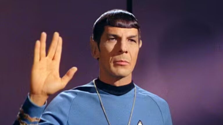 Personagem de orelhas pontudas e cabelo preto curto com franja reta usa uniforme azul com insígnia no peito e faz o gesto vulcano de saudação com a mão direita levantada e os dedos separados em V, sobre fundo roxo uniforme.