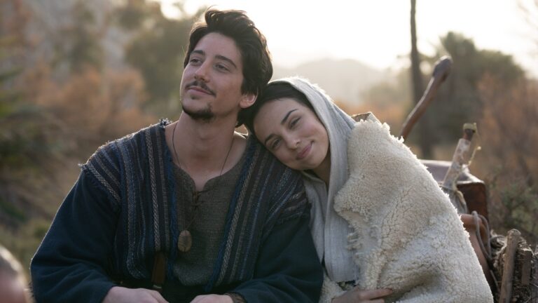 Casal com trajes de época senta ao ar livre em paisagem rural com a mulher apoiando a cabeça no ombro do homem e objetos de viagem ao lado