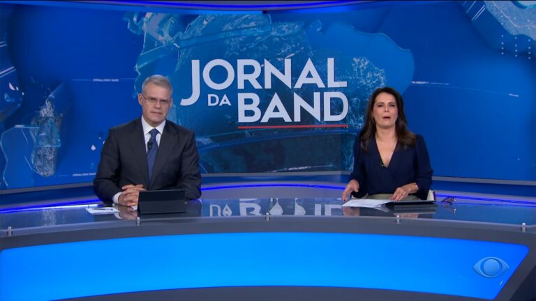 A imagem mostra Eduardo Oinegue e Adriana Araújo no estúdio do Jornal da Band, sentados atrás da bancada principal do telejornal. Eduardo Oinegue, à esquerda, veste terno escuro com gravata azul, enquanto Adriana Araújo, à direita, usa uma blusa azul-marinho. Atrás deles, um grande painel azul exibe o logotipo do Jornal da Band em letras brancas, compondo um cenário moderno e iluminado
