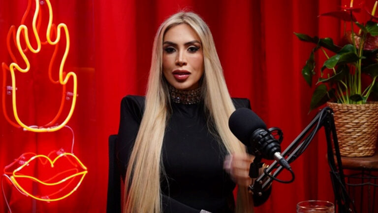 Juju Ferrari de preto e gargantilha fala ao microfone em estúdio com cortina vermelha e neons de fogo e boca iluminados atrás